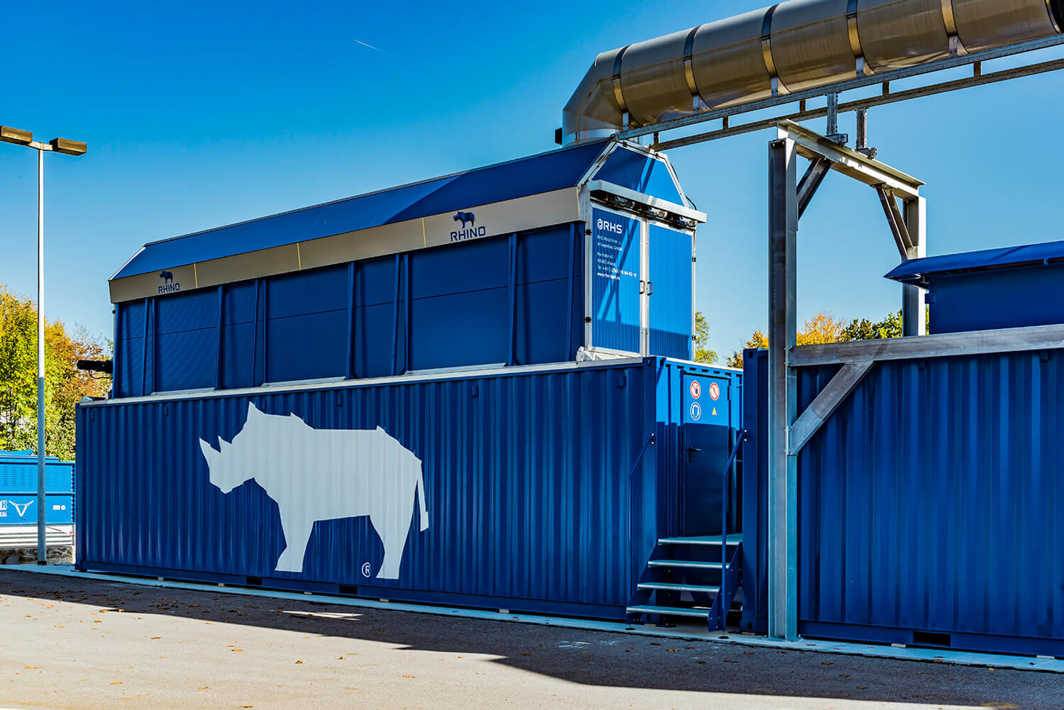 Ein großer blauer Schiffscontainer, auf dessen Seite die Silhouette eines weißen Nashorns gemalt ist, steht an einem sonnigen Tag im Freien und ist mit einem Metallrohrsystem verbunden.