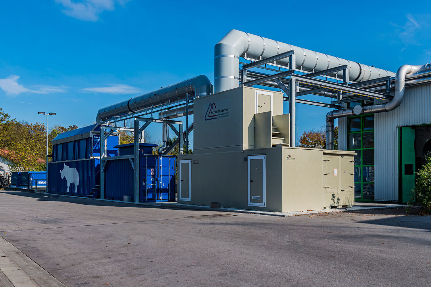 Eine Industrieanlage mit großen blauen und grauen Geräten, sichtbaren Rohren und einer Gebäudehülle. Auf blauen Containern sind die Silhouetten von Kühen und Schweinen zu sehen. Der Himmel ist klar und der Boden ist gepflastert.