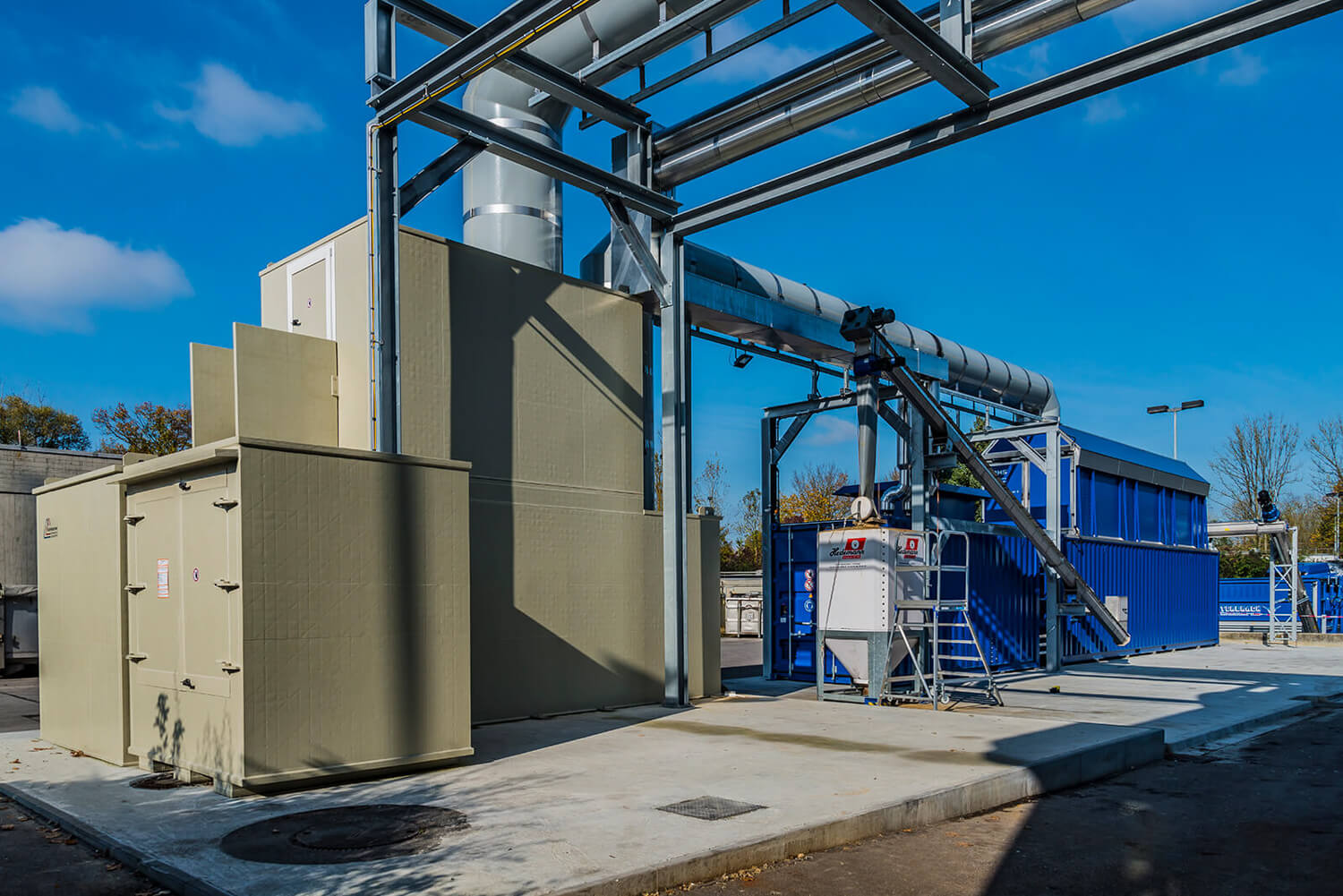 Eine Industrieanlage mit großen blauen und beigen Maschinen, die durch Metallrohre miteinander verbunden sind, steht im Freien auf einer Betonfläche unter strahlend blauem Himmel.