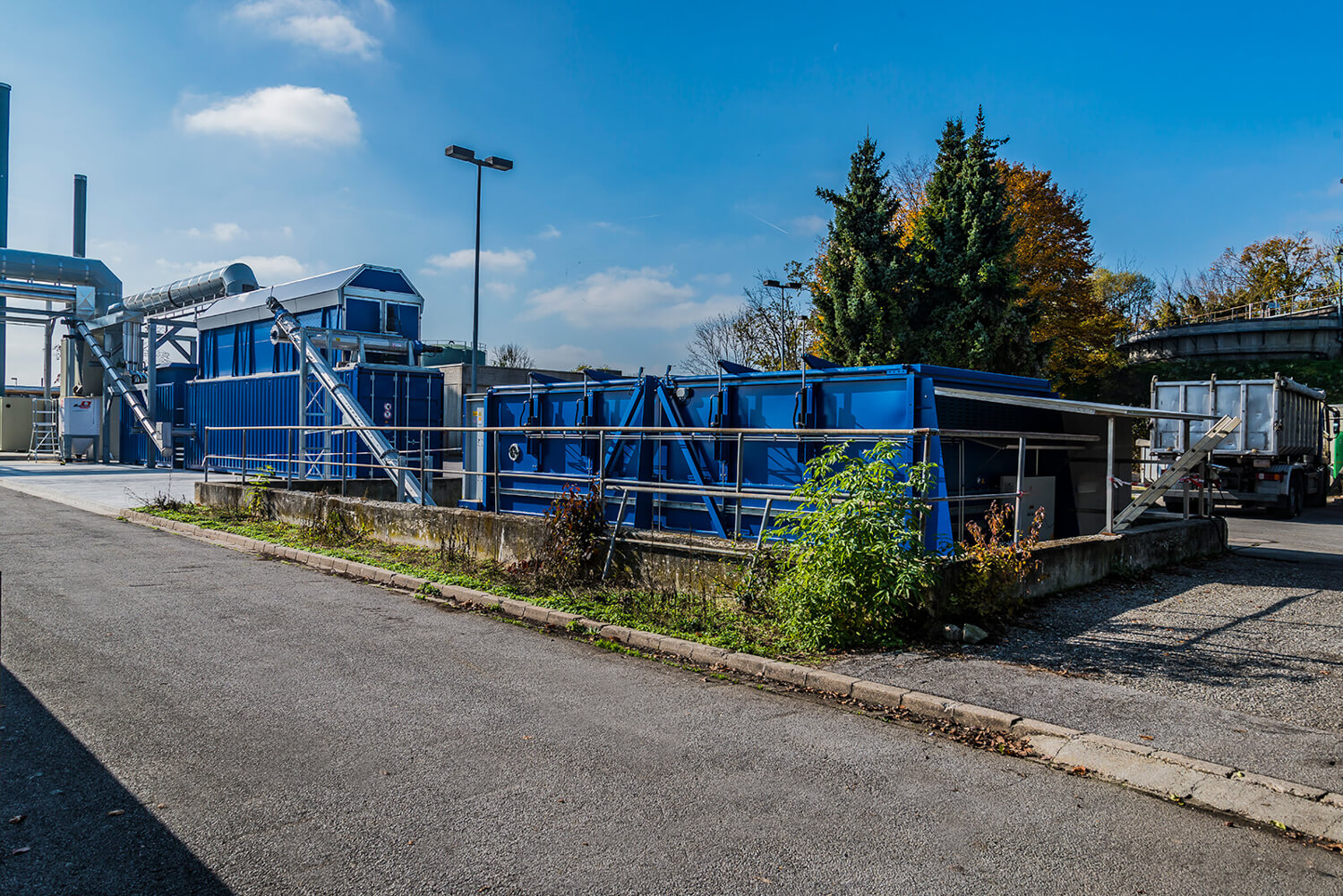 Industrieanlage mit mehreren großen blauen Metallcontainern und einem Gebäude. Im Hintergrund stehen Bäume und eine Straßenlaterne bei klarem Himmel. Die Vegetation wächst entlang des Bordsteins im Vordergrund.