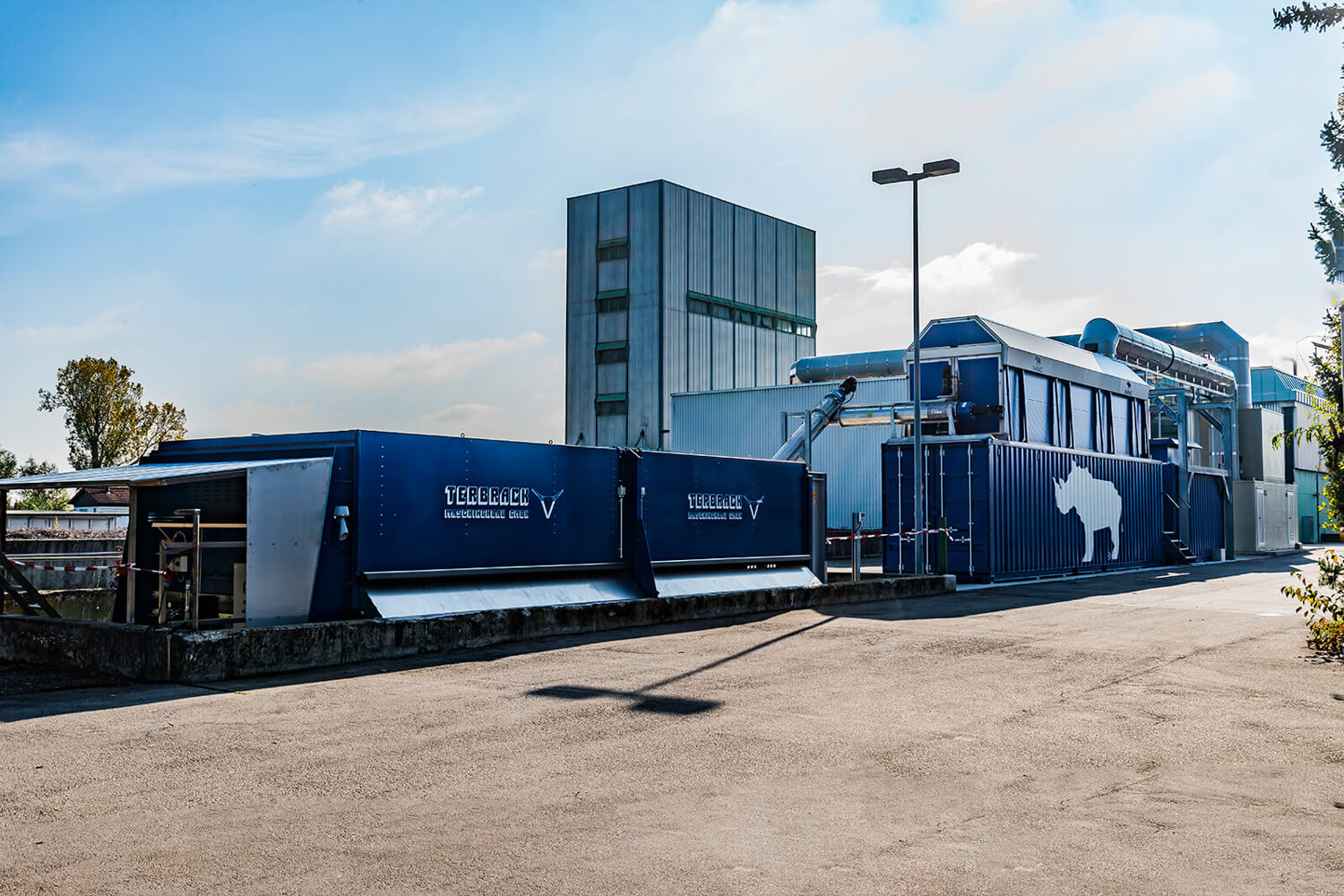 Eine moderne Industrieanlage mit blauen Containern mit der Aufschrift "TEGRASE" und dem Logo eines Nashorns neben einem hohen grauen Gebäude unter einem teilweise bewölkten Himmel.