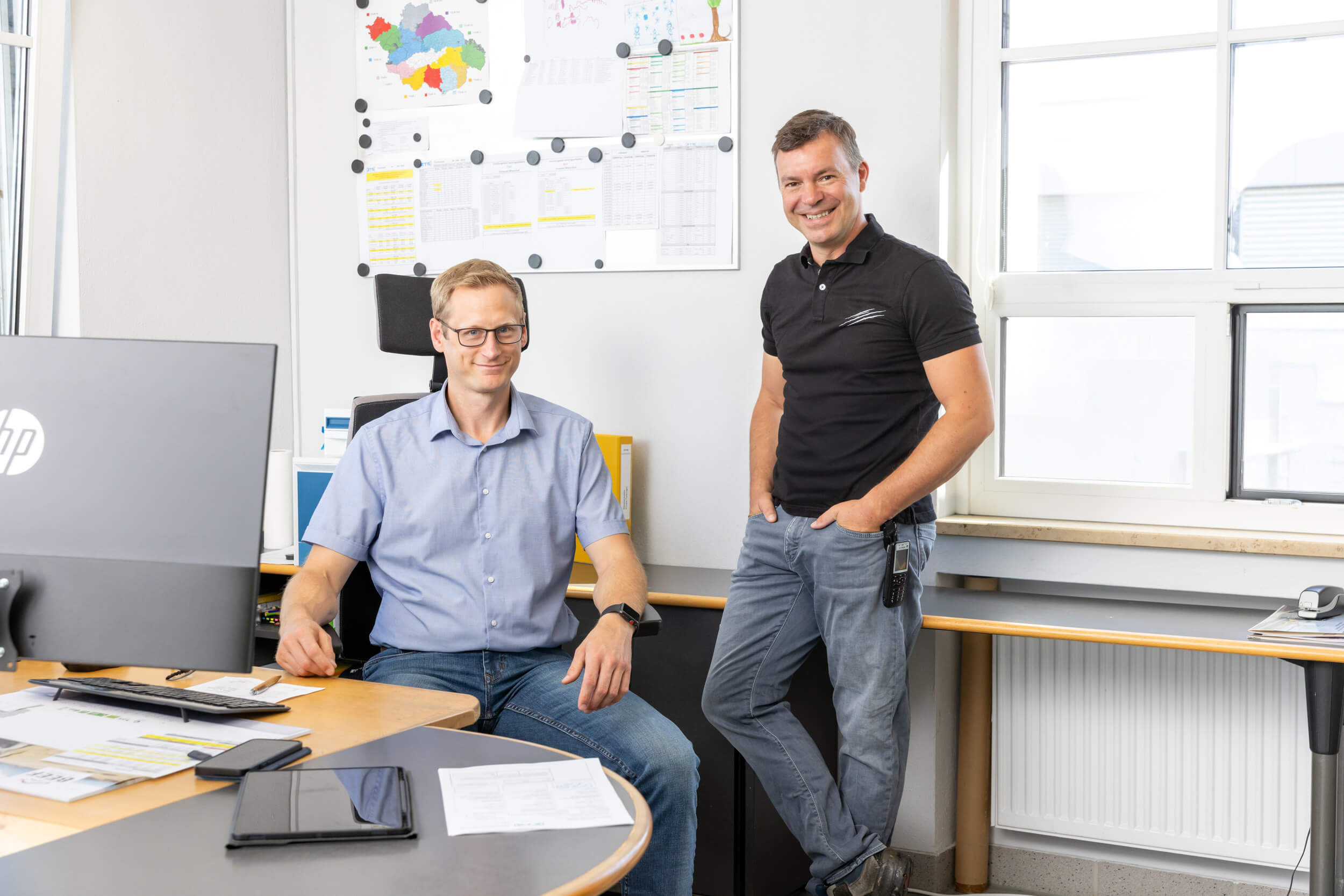 Zwei Männer in einem Büro: Der eine sitzt an einem Schreibtisch mit Papieren, einem Monitor und einem Tablet, der andere steht daneben, beide lächeln. Hinter ihnen befinden sich ein Fenster und eine Wand mit Tabellen und Karten.
