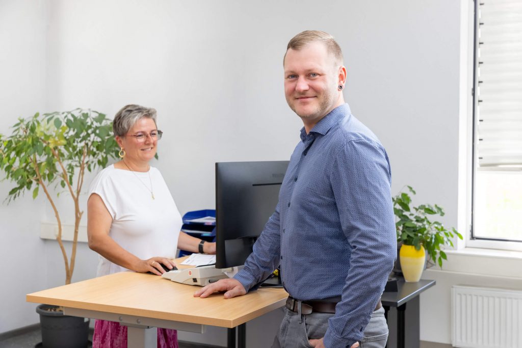 Ein Mann in einem blauen Hemd steht in einem Büro und lächelt in die Kamera, während eine Frau in einem weißen Oberteil an einem Stehpult im Hintergrund arbeitet.