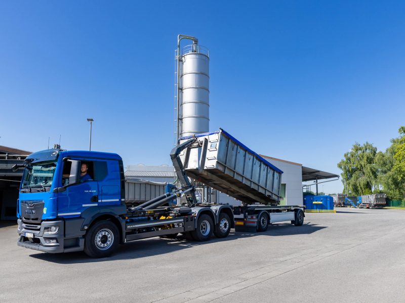 Ein blauer Lastwagen mit einem Kippanhänger entlädt unter blauem Himmel Fracht in einer Industrieanlage. Im Hintergrund sind ein hohes Metallsilo und verschiedene Gebäude zu sehen.