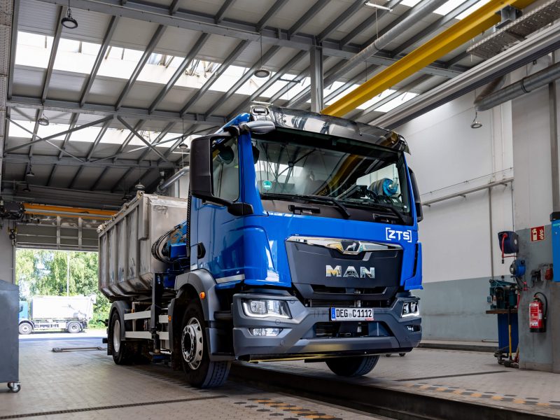 Ein blauer MAN-Lkw mit einem Frachtcontainer ist in einer großen, gut beleuchteten Industriegarage oder Werkstatt geparkt, in der im Hintergrund Geräte und Werkzeuge zu sehen sind.