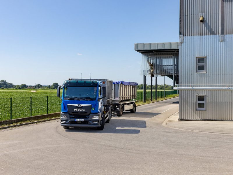 Ein blauer MAN-Lkw mit Anhänger verlässt ein Industriegebäude und fährt auf eine asphaltierte Straße. Im Hintergrund sind grüne Felder und ein blauer Himmel zu sehen.