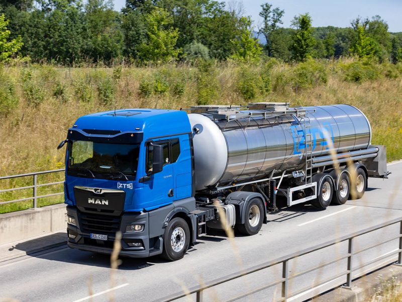 Ein blauer Tankwagen mit einem silbernen ZTS-Tankanhänger fährt an einem sonnigen Tag auf einer gepflasterten Straße, die von Gras und Bäumen gesäumt ist.
